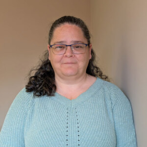 A woman with long dark curly hair and glasses wearing a blue sweater.