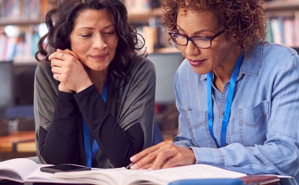 Two women wearing blue lanyards around their neck are sitting at a desk looking through a book.