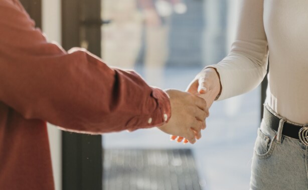 A person in a brown shirt is shaking hands with a woman in a cream coloured top and light jeans