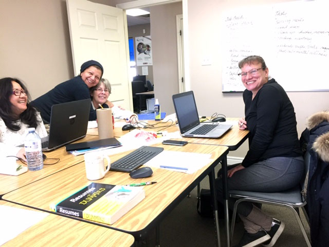 Four smiling women sit around a group of desks with laptop computers open. There is a white board with writing on it beside them.