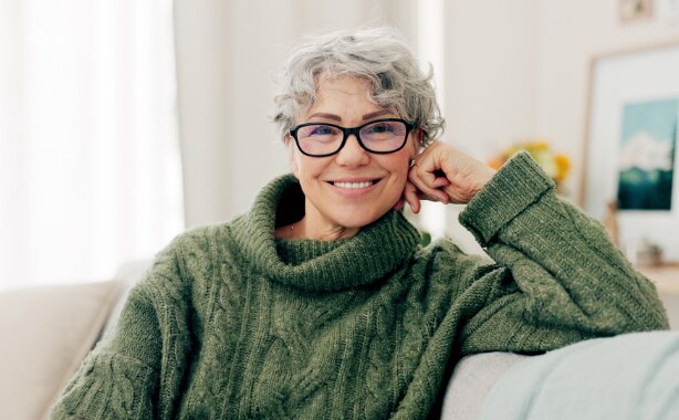 A middle-aged woman in a green sweater with short curly grey hair and glasses sitting on a couch