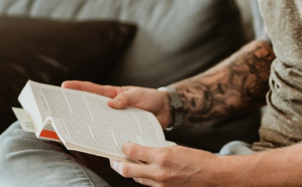 A man with tattoos on his left arm is sitting on a couch reading a book