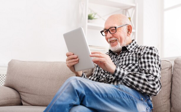 An older man smiles as he sits on a couch looking at something on his tablet