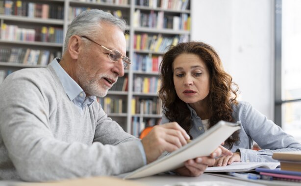 An older man with grey hair and beard points to an open book while a woman with long dark hair looks on