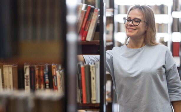 A woman in a white shirt and glasses is reaching for a book from a black shelf containing multiple books