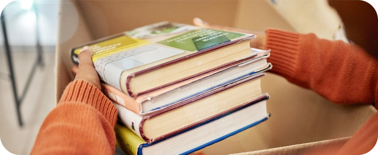 Close up of the arms of a person in an orange sweater pulling 4 used books out of a box